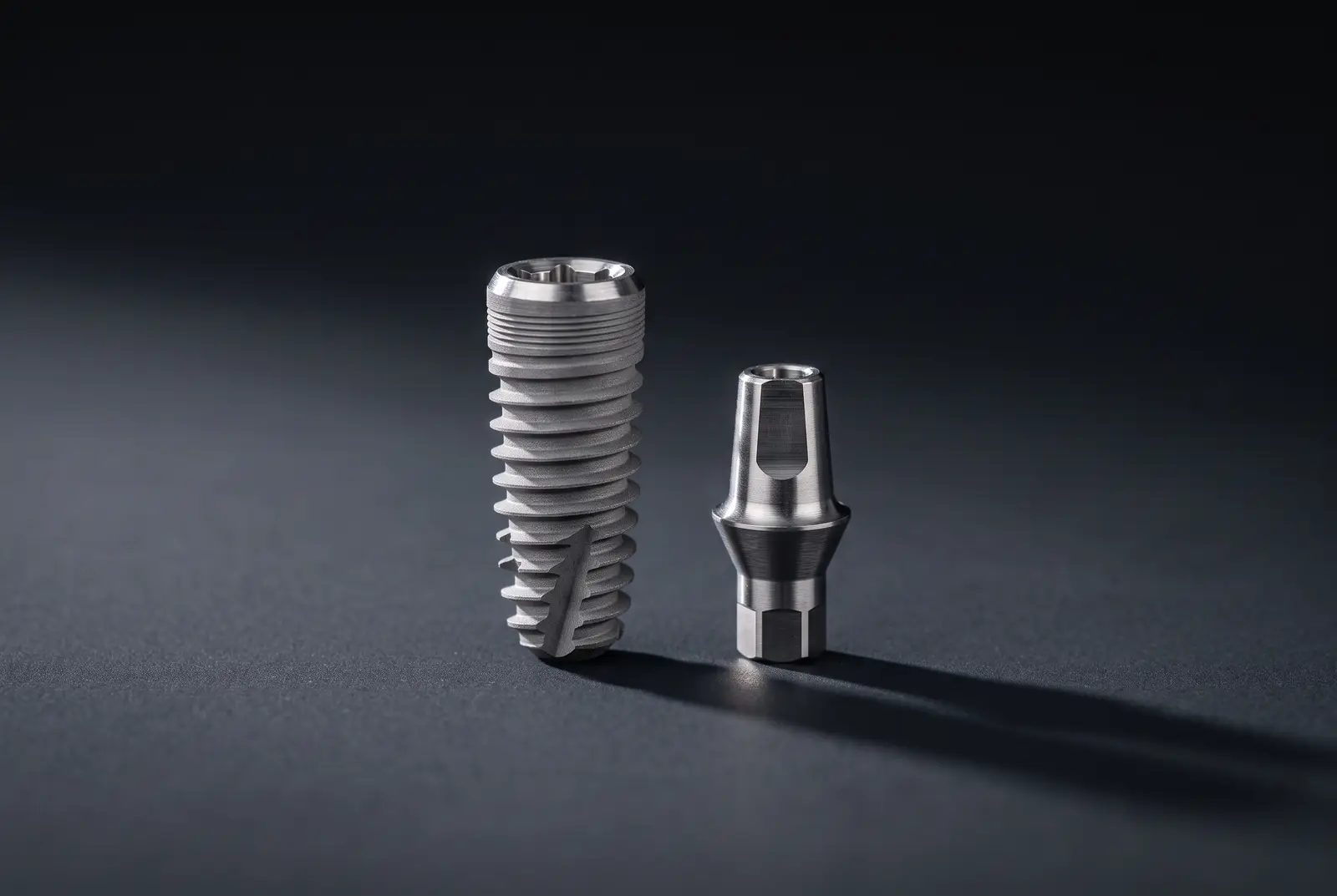 A titanium implant post with a machined thread, standing beside the smaller abutment that connects it to a crown.